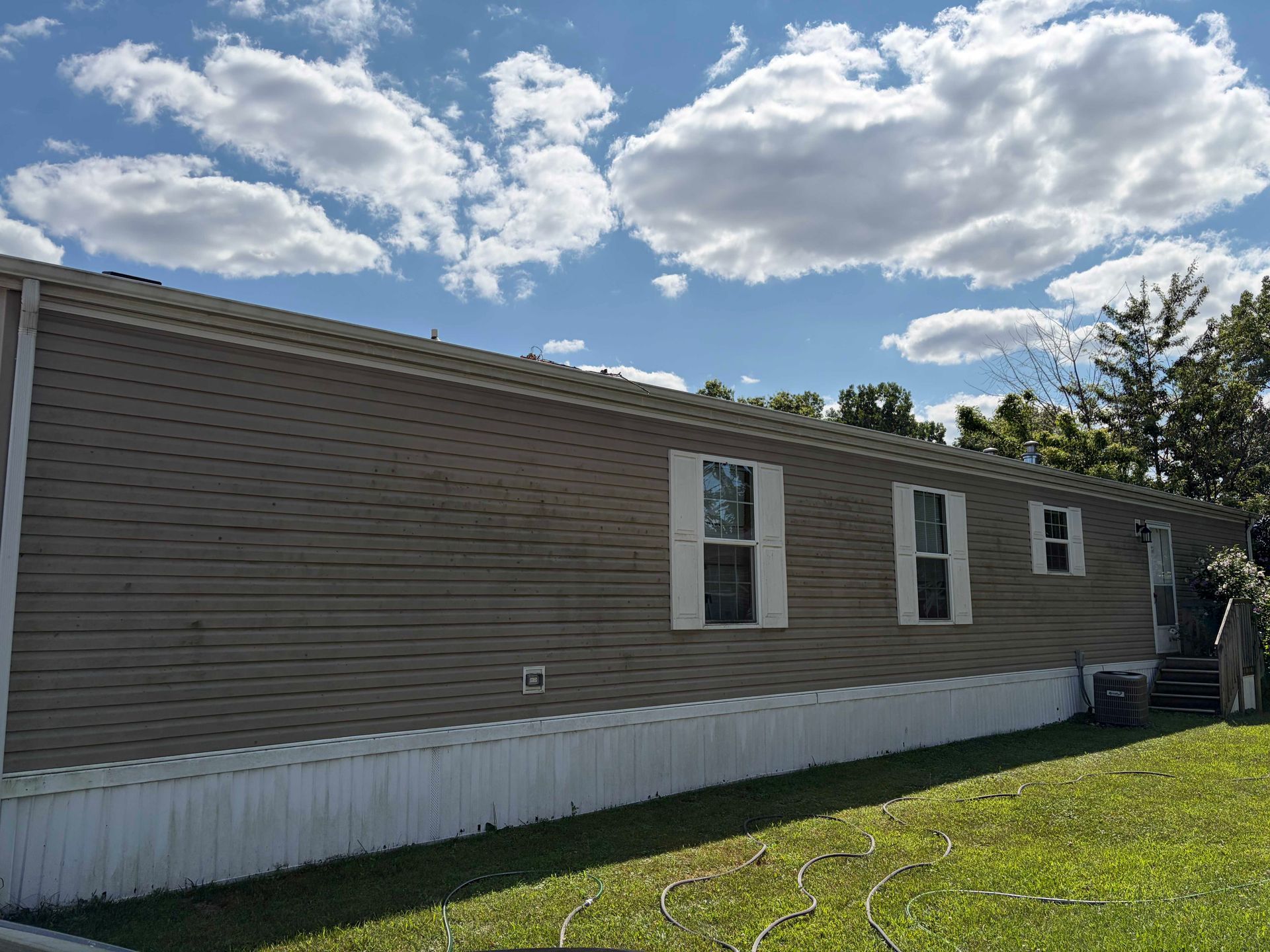 Single-story beige mobile home with white trim under a partly cloudy sky, beside a grassy lawn.