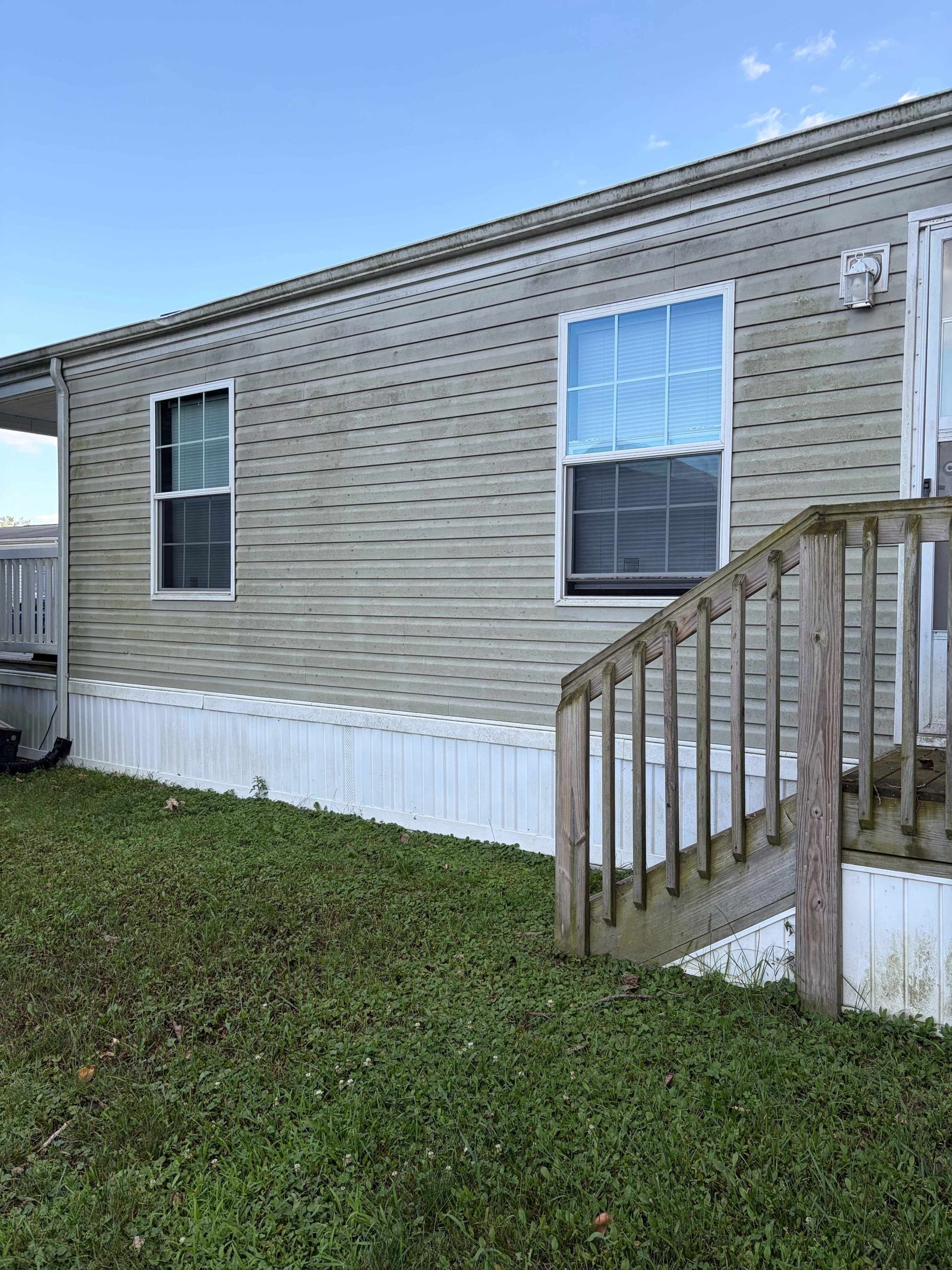 Mobile home exterior with beige siding, white skirting, and a wooden porch railing by a grassy yard