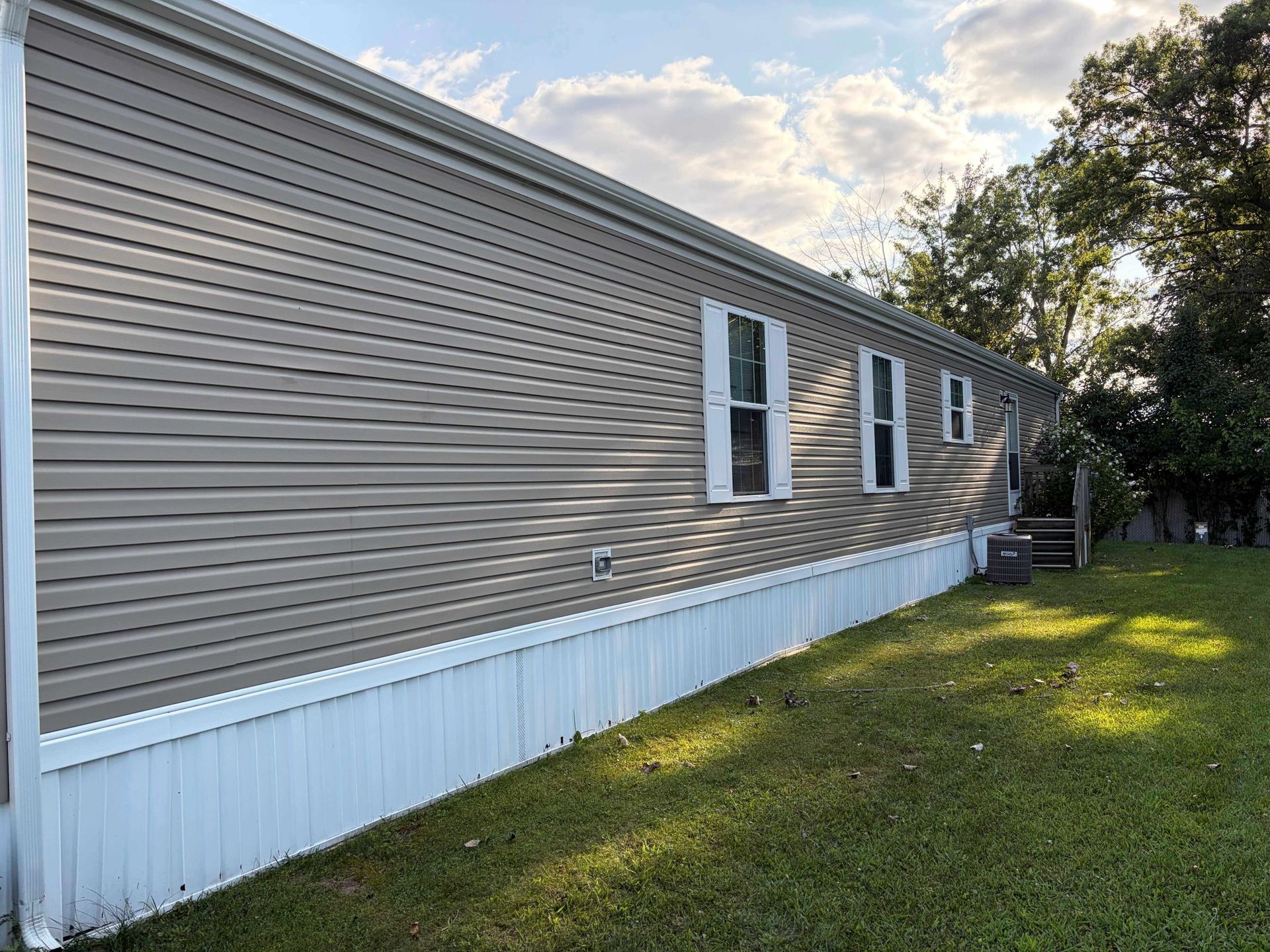Side view of a beige mobile home with white skirting, windows, and a grassy yard under a partly cloudy sky