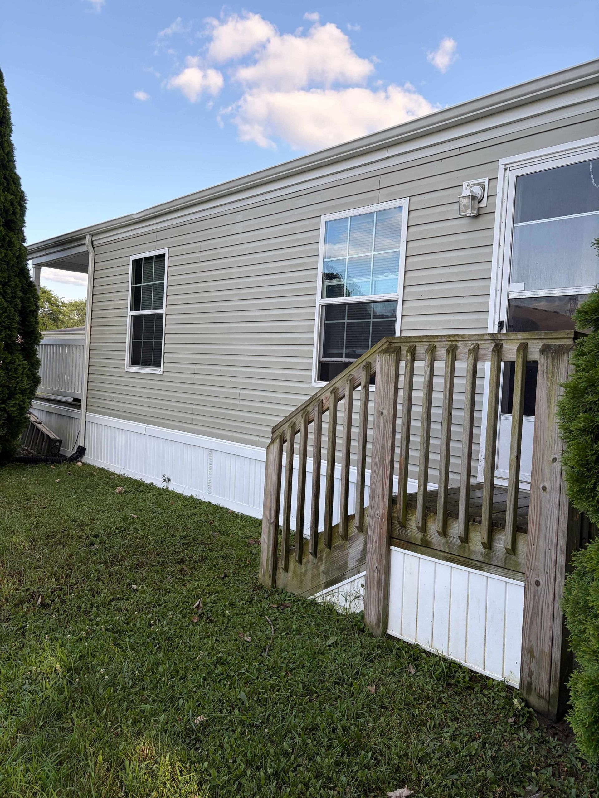 Mobile home exterior with beige siding, white skirting, and a small wooden ramp on a grassy lot