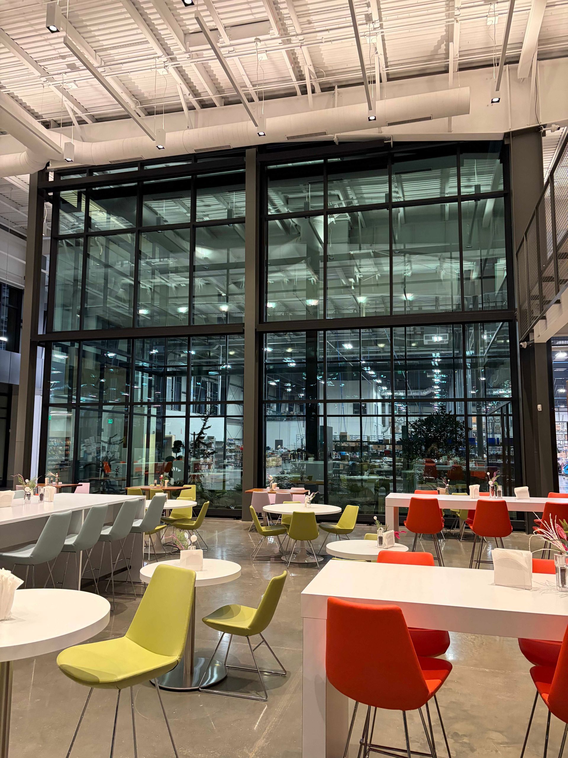Modern glass-walled cafe with white tables and colorful chairs under a high industrial ceiling
