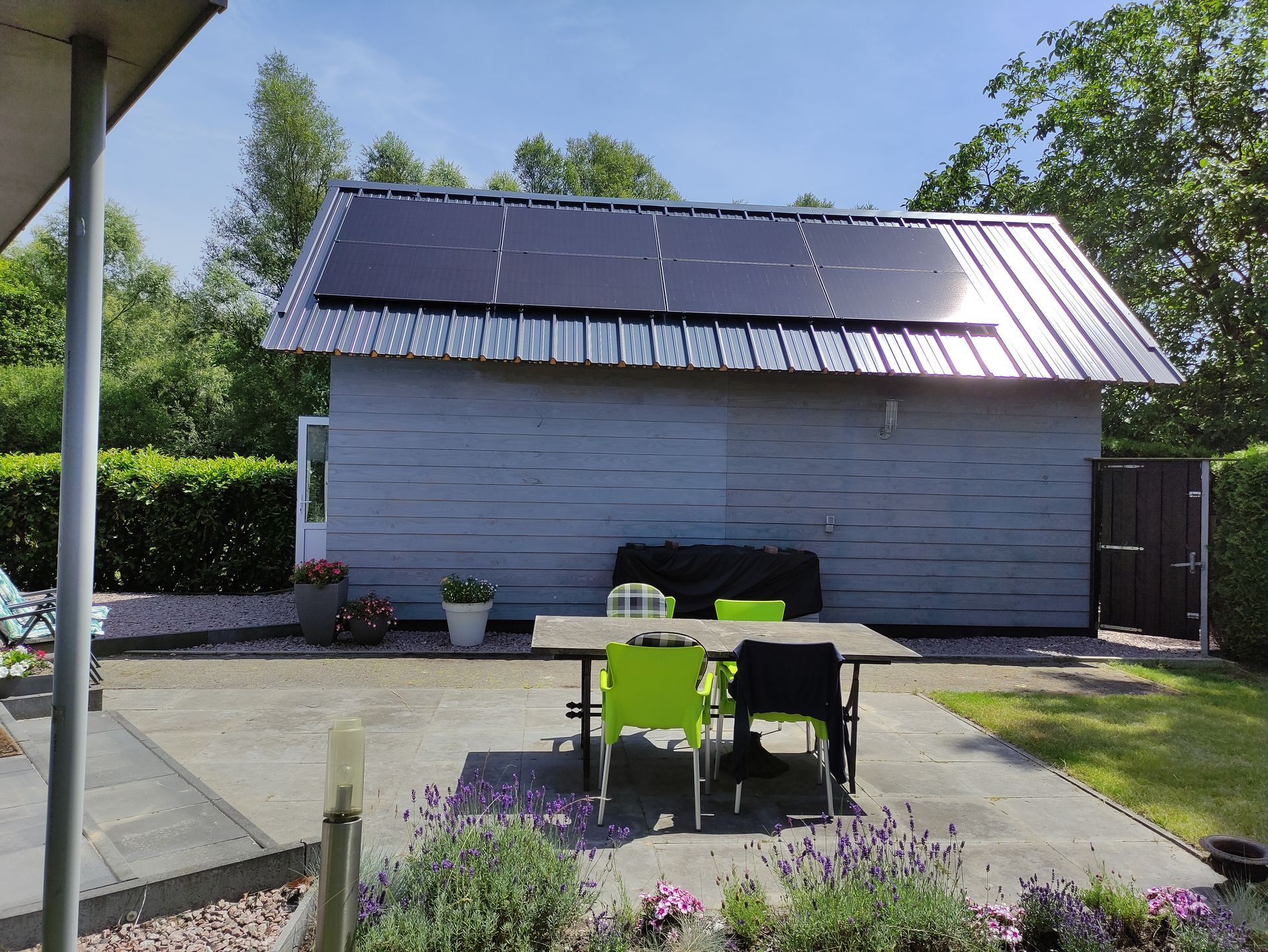 Terras in de achtertuin met tafel en stoelen tegenover een schuurachtig gebouw met zonnepanelen op het dak.
