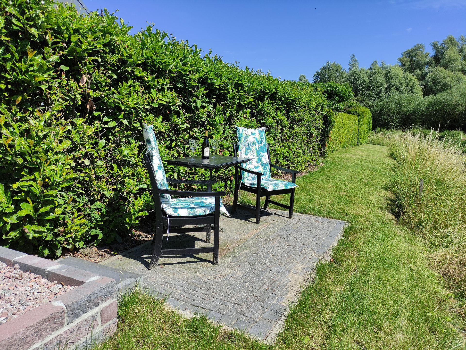 Twee zwarte tuinstoelen op een grindpad naast een groene heg en gras.