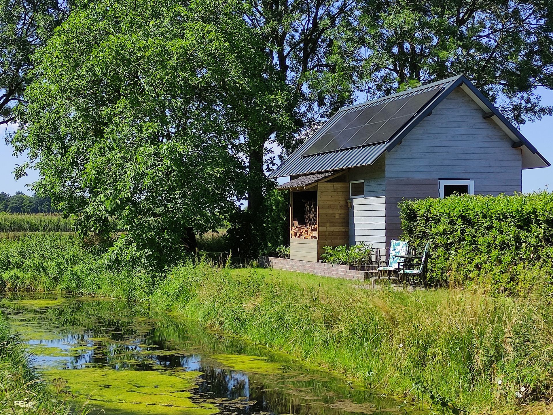 Klein grijs huisje aan een vijver, omgeven door groen gras en bomen in het heldere zonlicht.