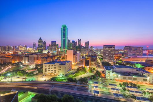 Dallas skyline at dusk, showcasing numerous illuminated skyscrapers under a purple and blue sky.