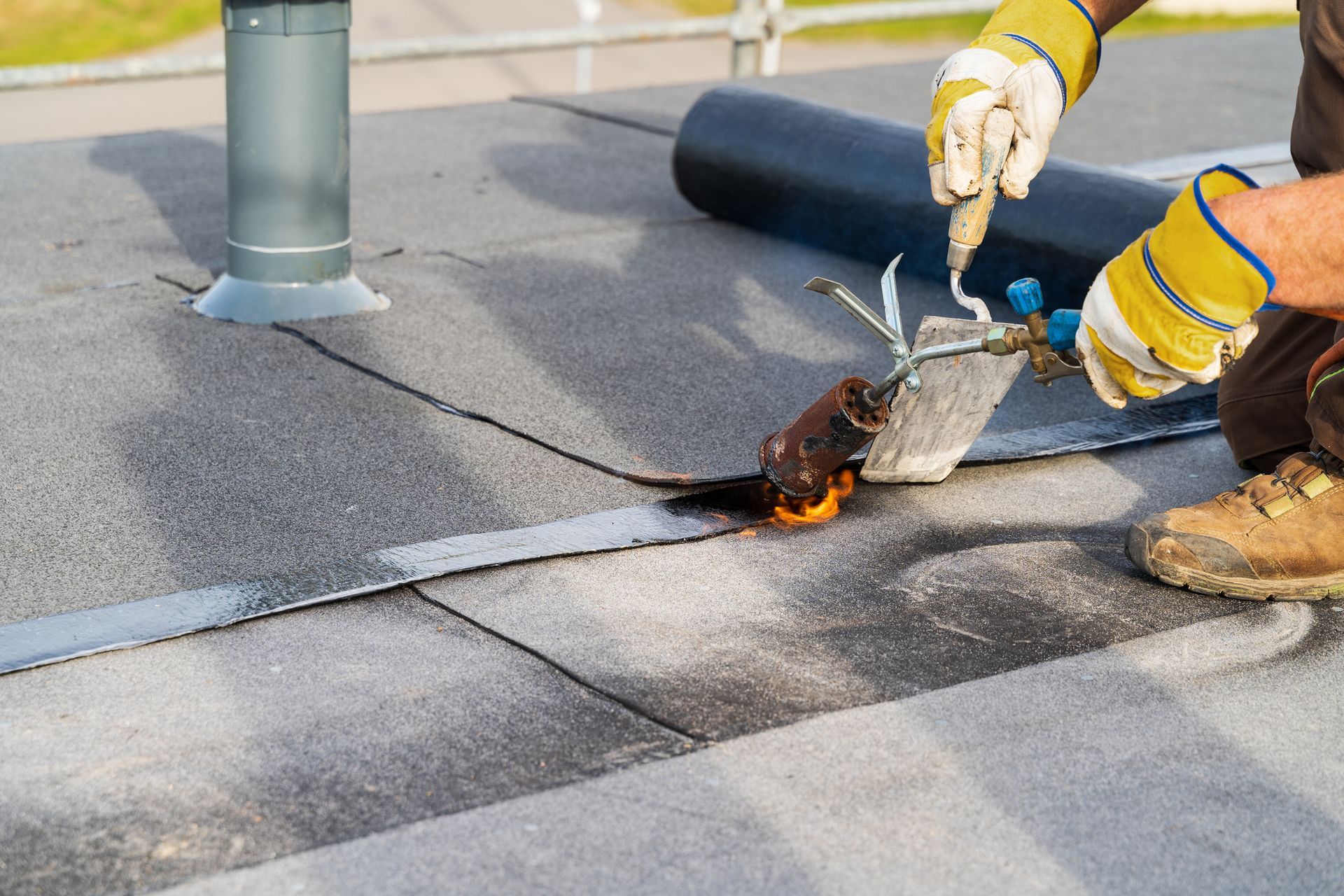 Person using a torch to seal seams on a flat roof, wearing gloves and safety boots.