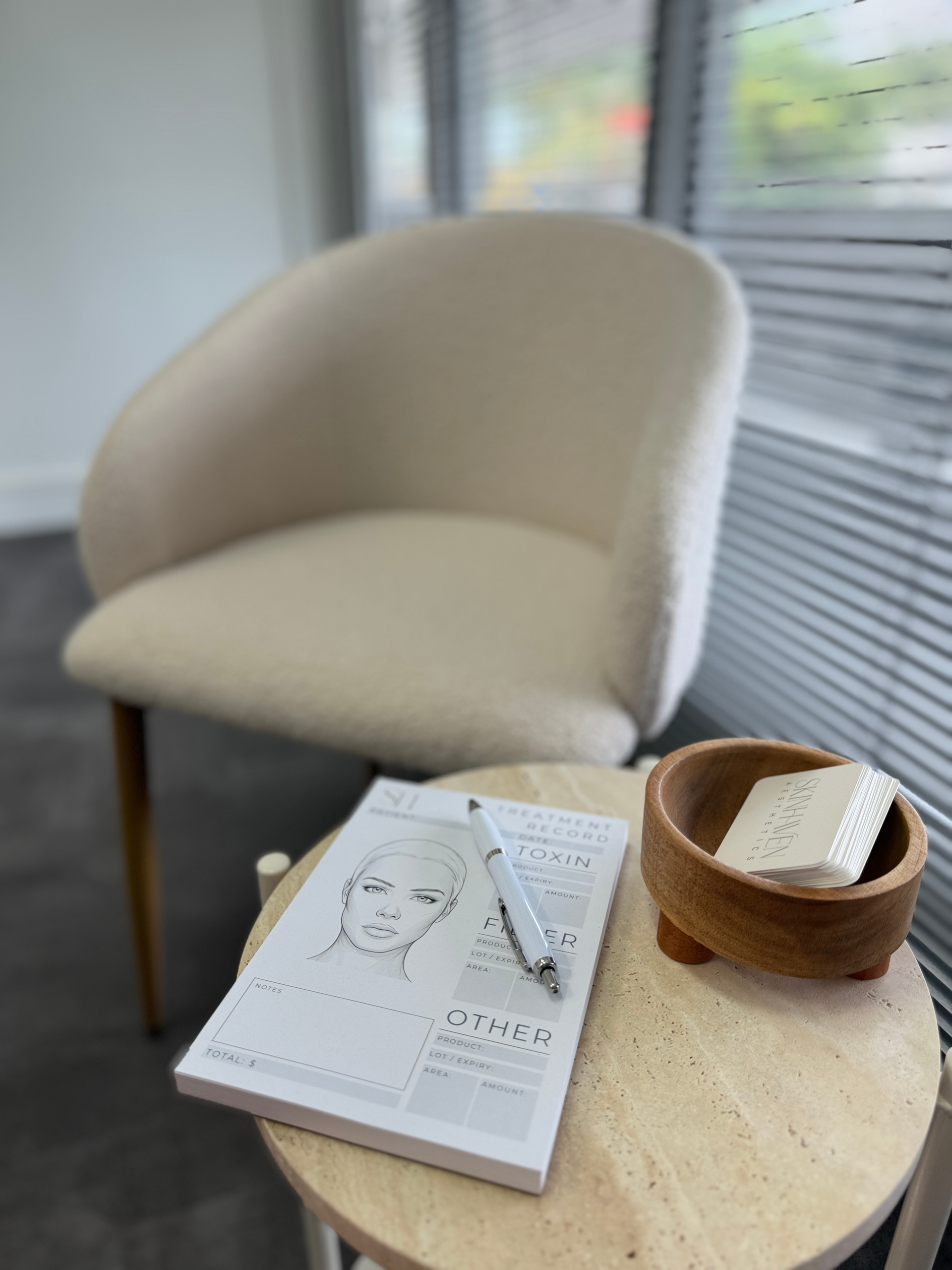 A beige armchair, a notepad with a facial diagram and a pen, and a small wooden bowl on a round stone side table — Skin Haven Aesthetics Whitsundays in Airlie Beach, QLD