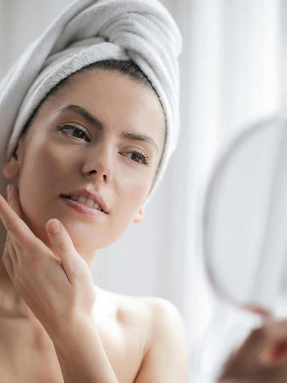 A person wearing a white head towel, touching their face while looking into a handheld mirror — Skin Haven Aesthetics Whitsundays in Airlie Beach, QLD