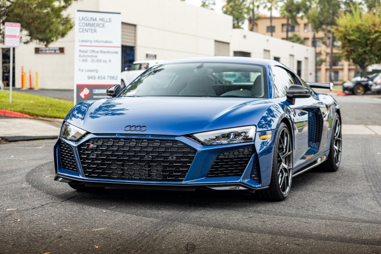 Blue Audi sports car parked outdoors at a car show, front view with city buildings in the backgroundSilver and neon yellow sports car with oversized rear wing parked beside a white wall. | Pacific German