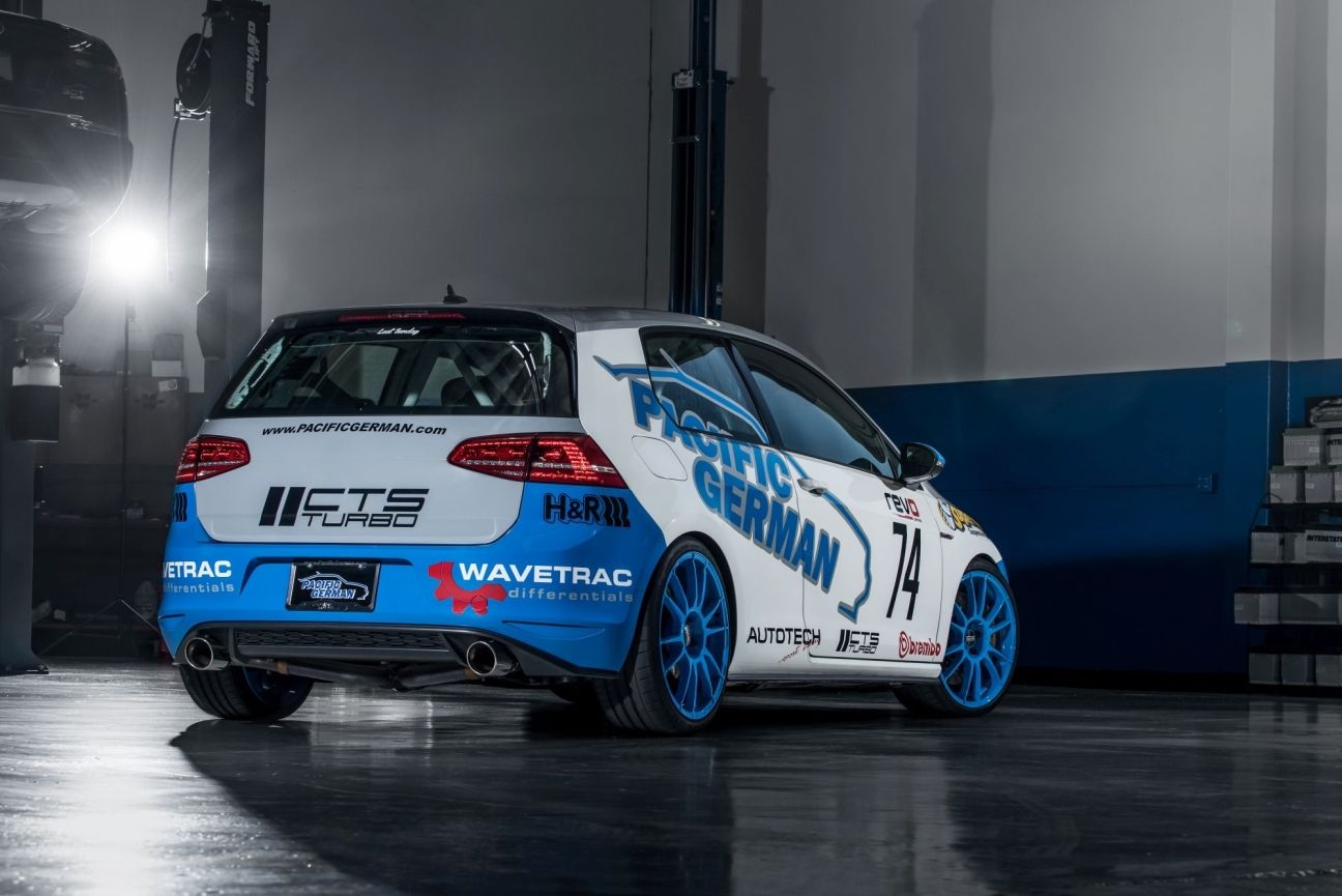 White-and-blue rally car in a garage, shown from the rear with sponsor decals and wet floor reflectionsSilver and neon yellow sports car with oversized rear wing parked beside a white wall. | Pacific German