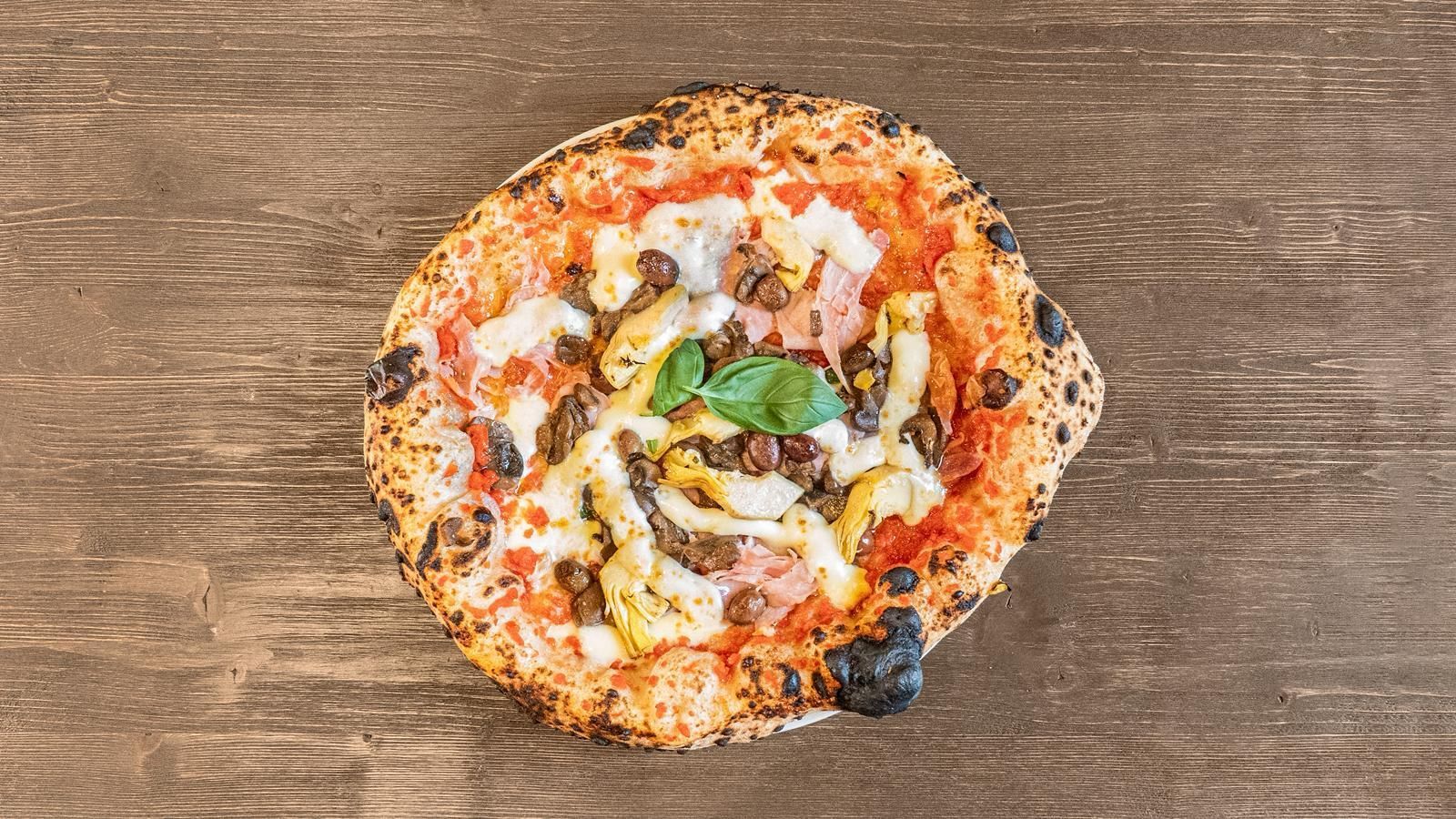 Vista dall’alto di una piccola pizza con basilico, funghi e formaggio fuso su un tavolo di legno.