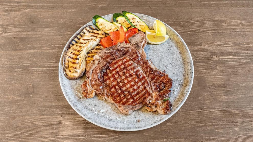 Bistecca alla griglia con gamberi, verdure e spicchi di limone su un piatto bianco.