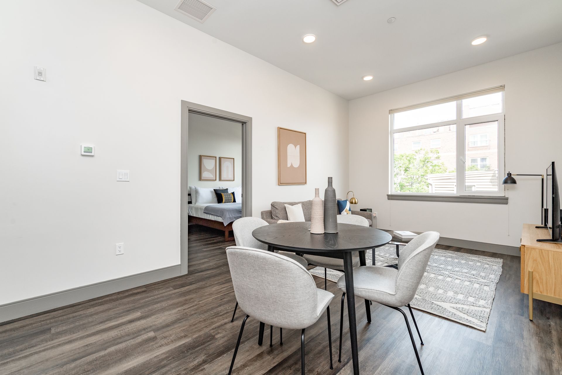 An open-concept living space with dining table, sofa, and window; bedroom visible in the background.