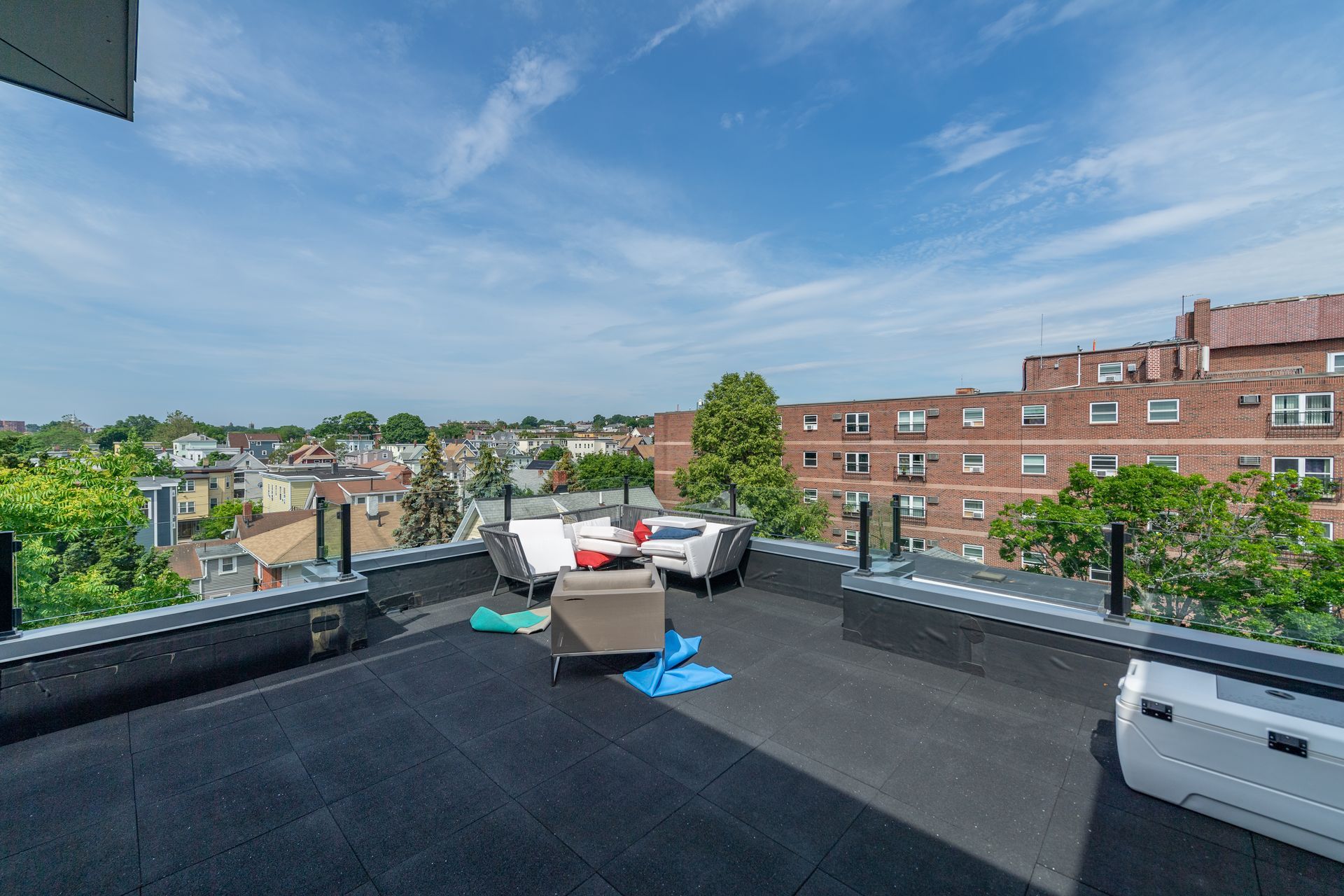 Rooftop patio with seating, cityscape view. Blue sky, brick buildings, trees. Black deck.