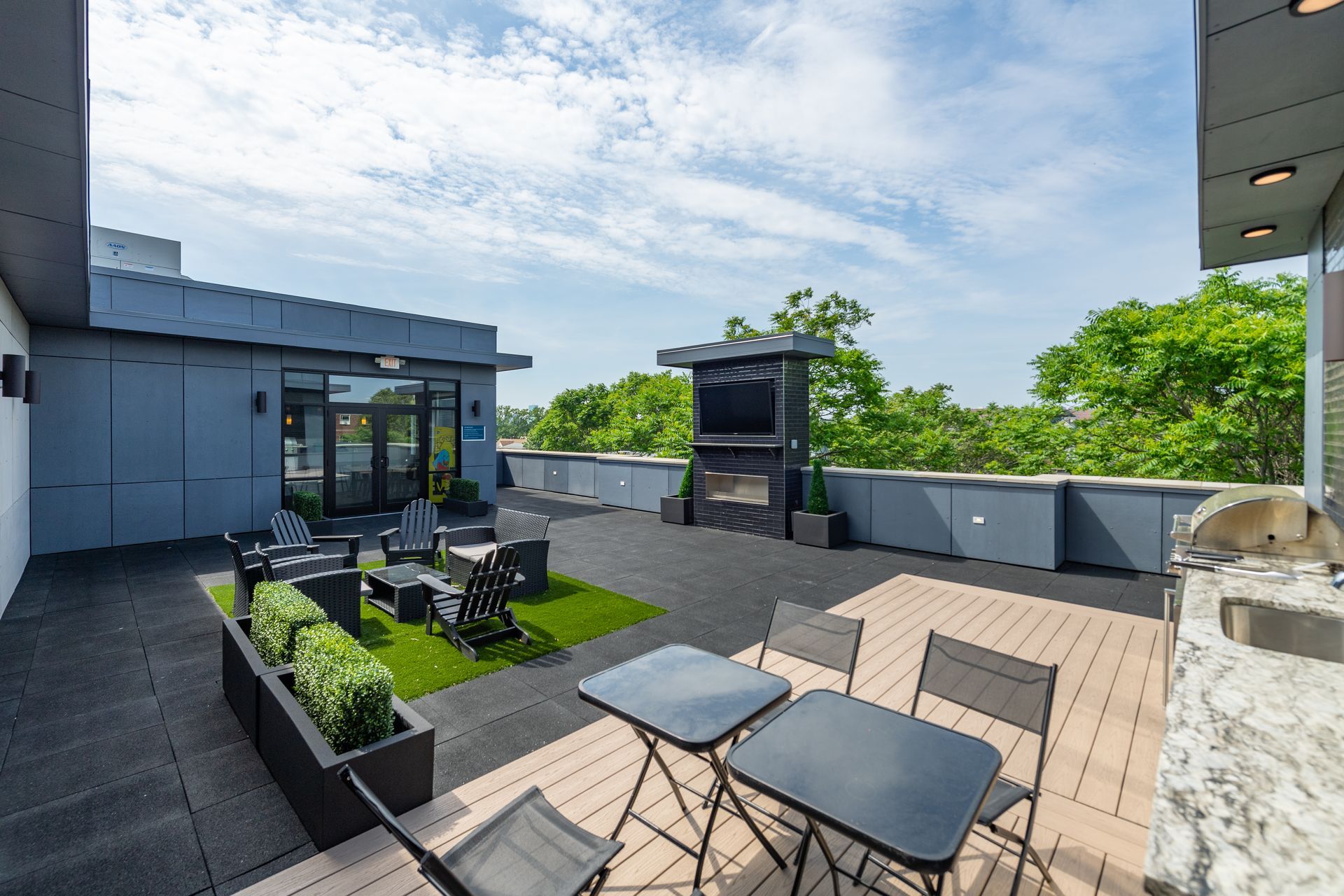 Rooftop patio with seating, fireplace, greenery, and partial city view under a blue sky.
