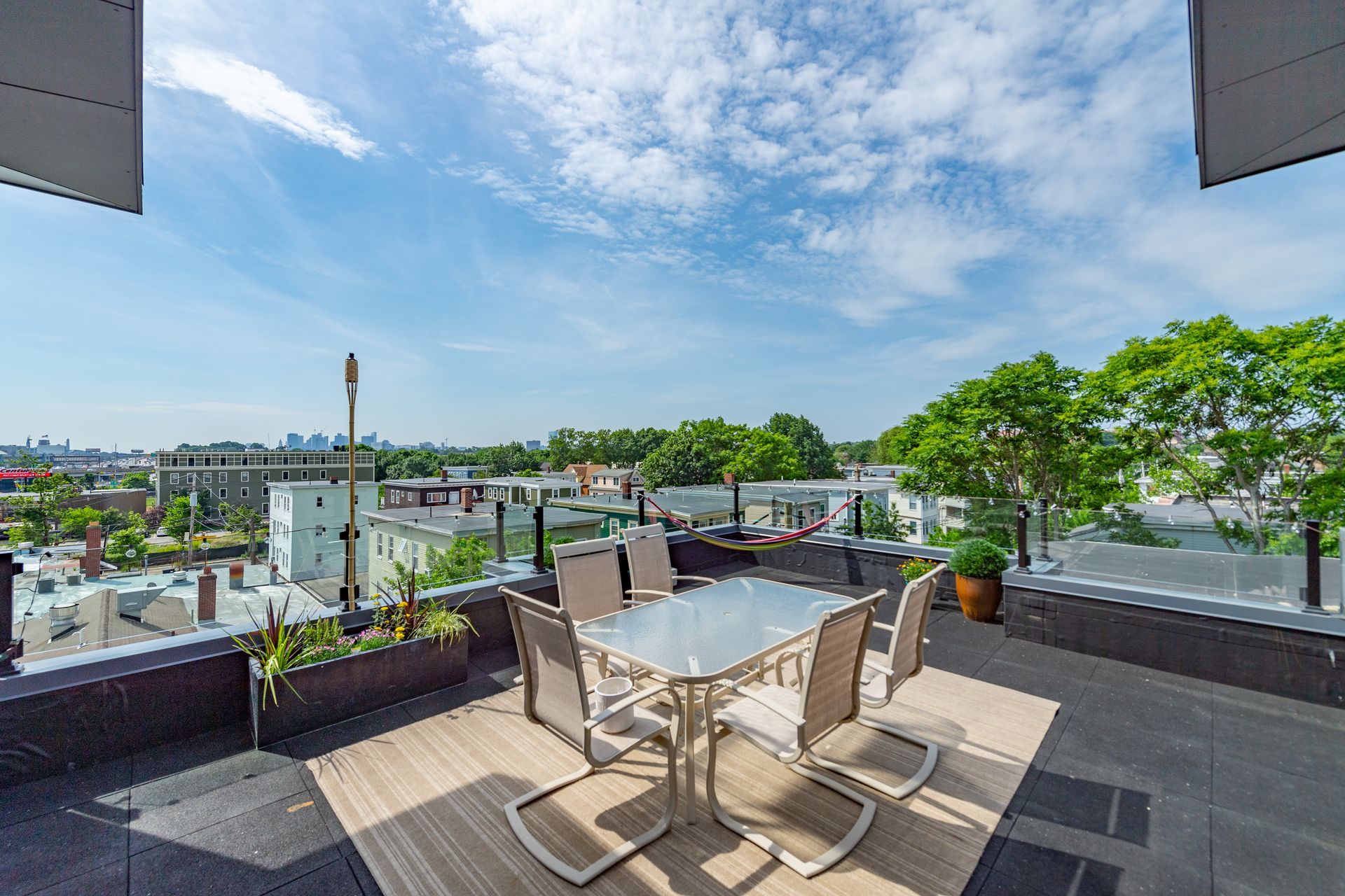 Rooftop patio with table, chairs, plants, and city view under a blue sky with clouds.