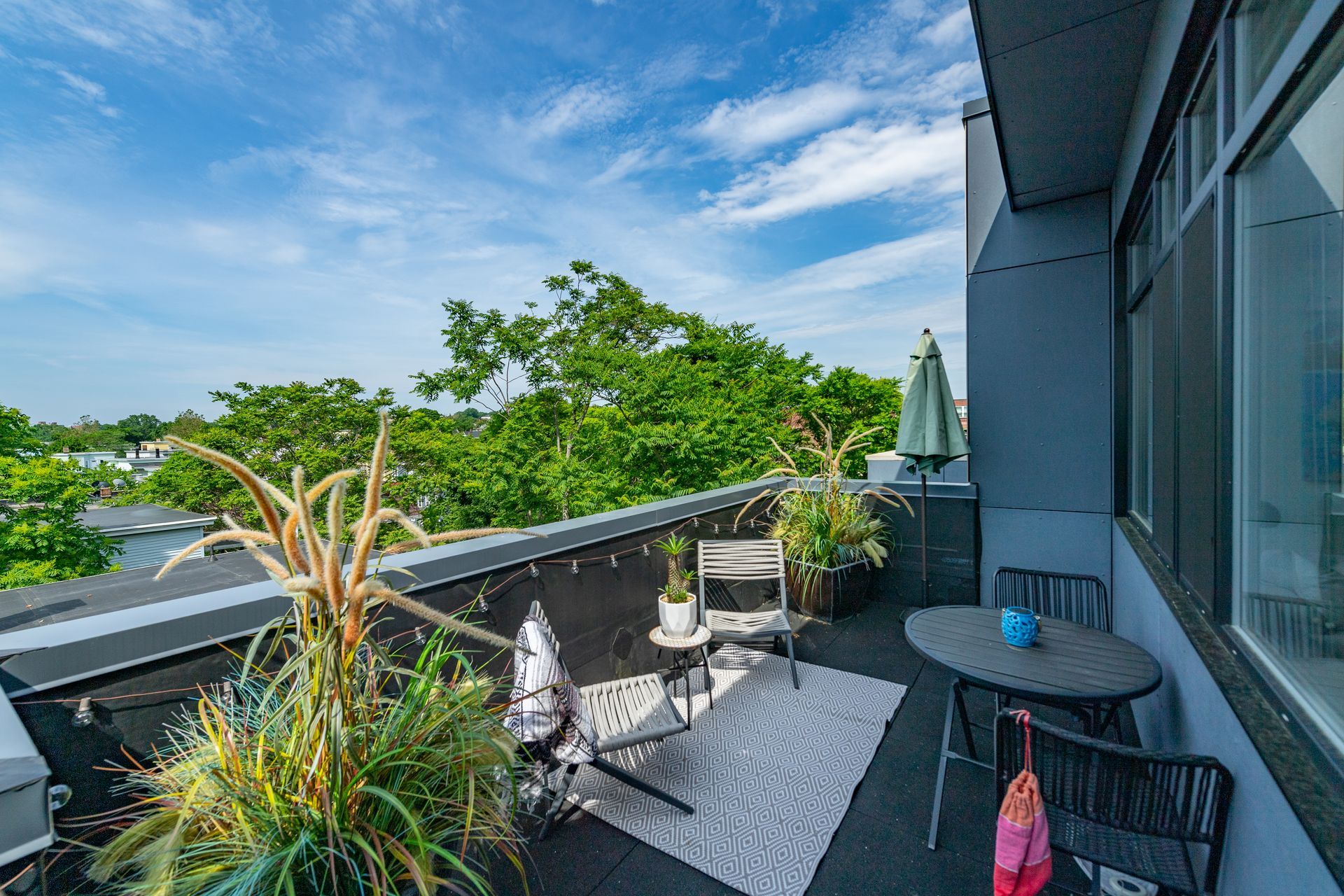 Rooftop patio with seating, plants, and umbrella. Blue sky and trees in the background.
