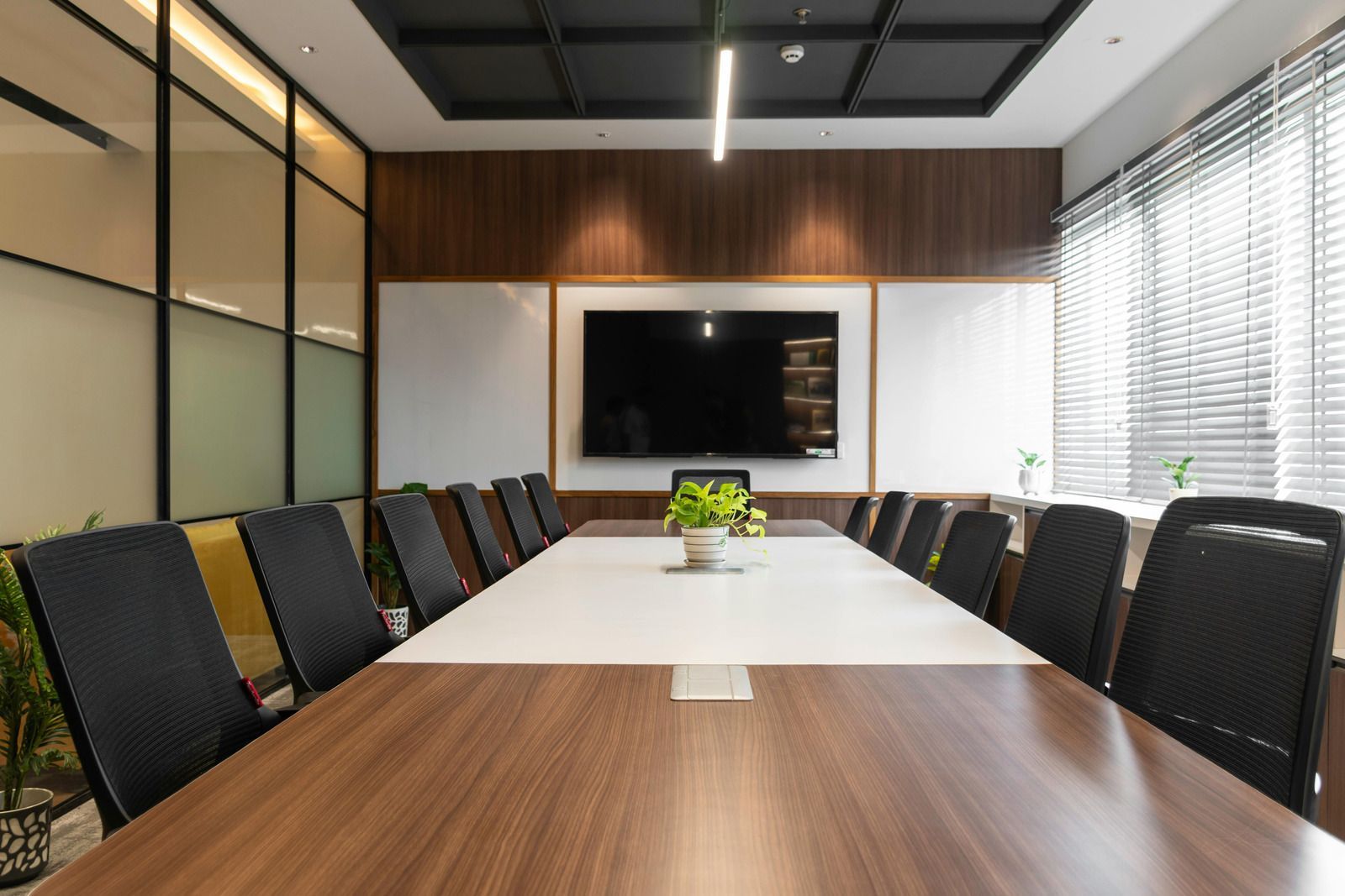 Modern conference room with long wooden table, black chairs, wall-mounted TV, and bright window blinds