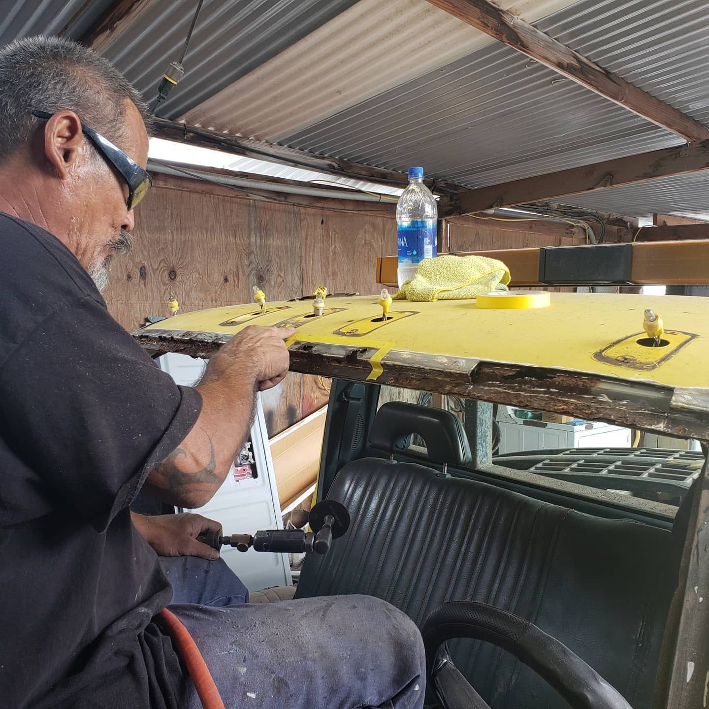 Person painting a yellow vehicle roof in a workshop, with a water bottle on top. | Rev Limit Auto Center