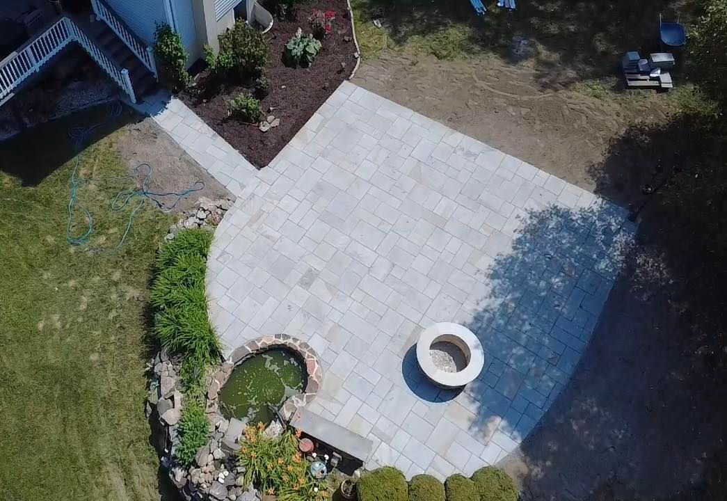 Aerial view of a patio with a fire pit, pond, and walkway in a backyard.