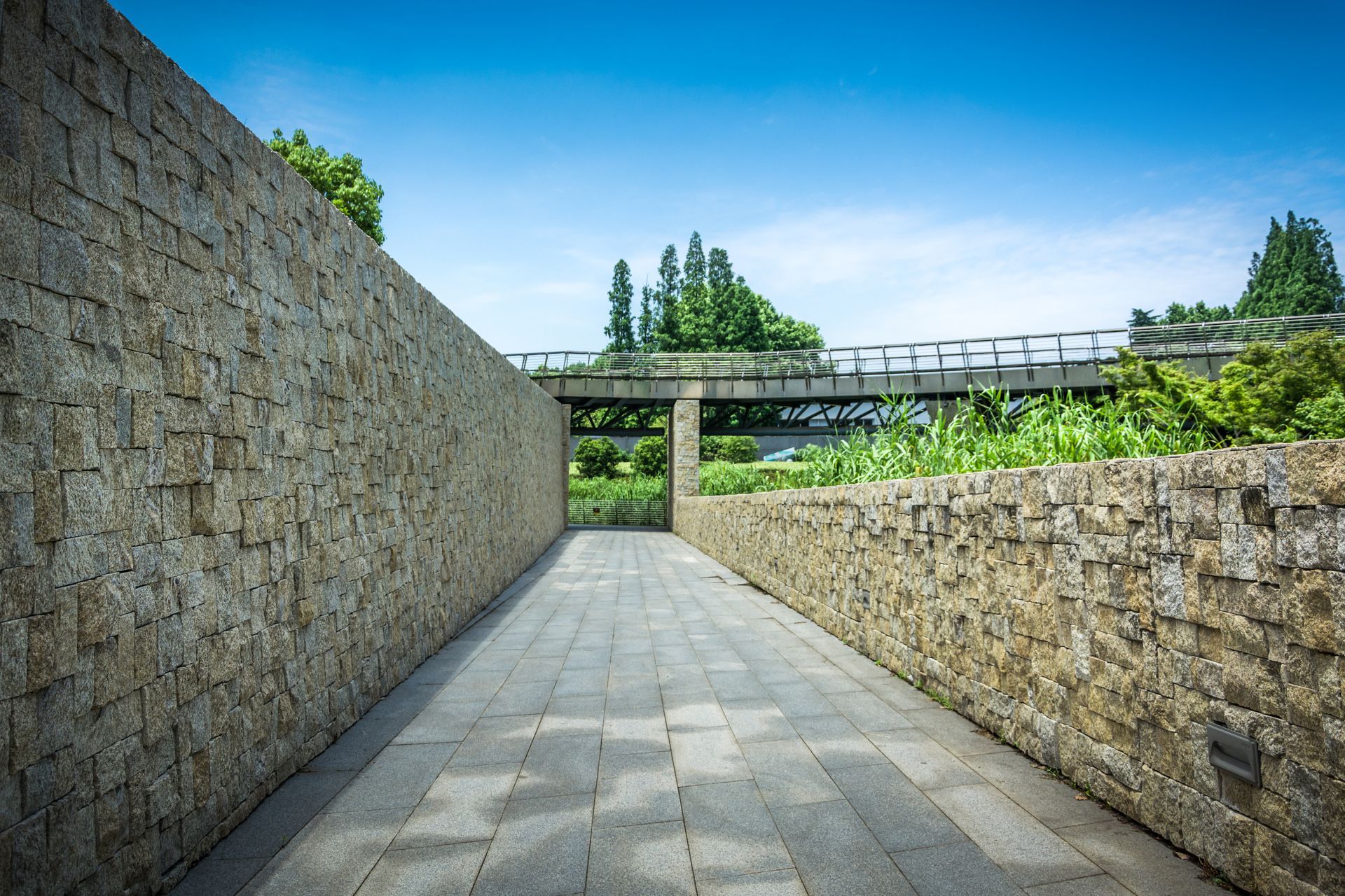 Stone pathway between high stone walls leading to a bridge and greenery under a blue sky.