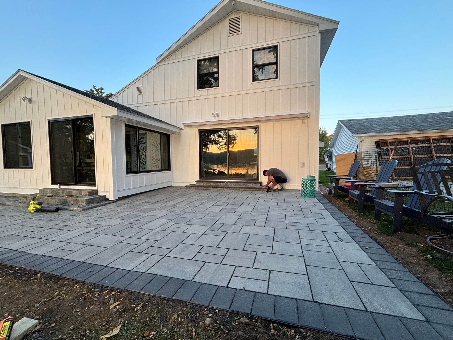 Backyard patio with gray pavers and a white house, two people working, and a view of water.
