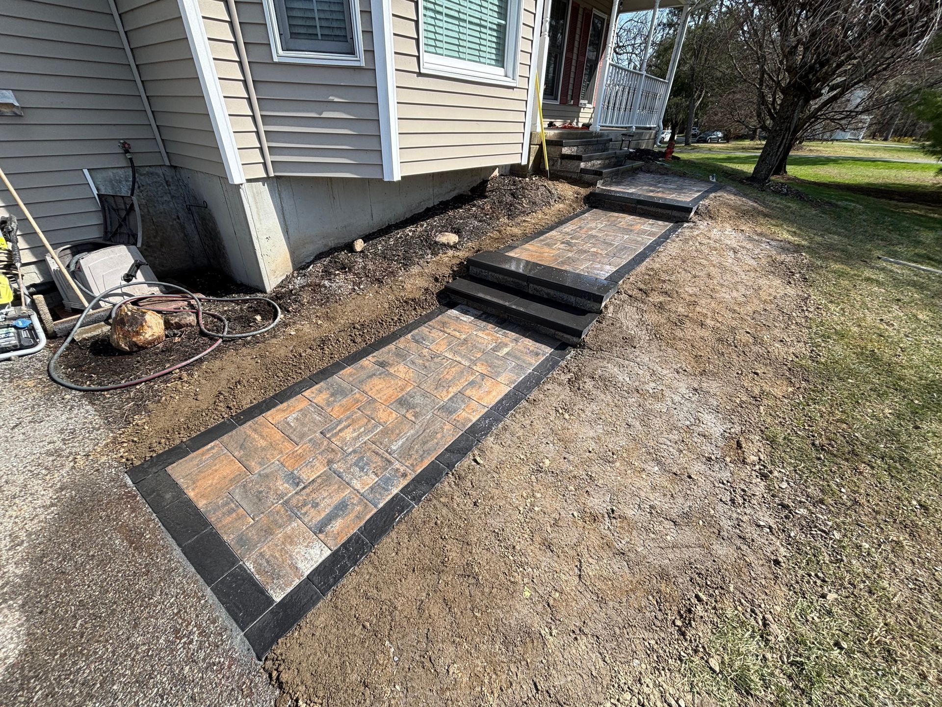 Brick pathway with steps leading up to a house entrance. Brown and black pavers set in a grassy yard.