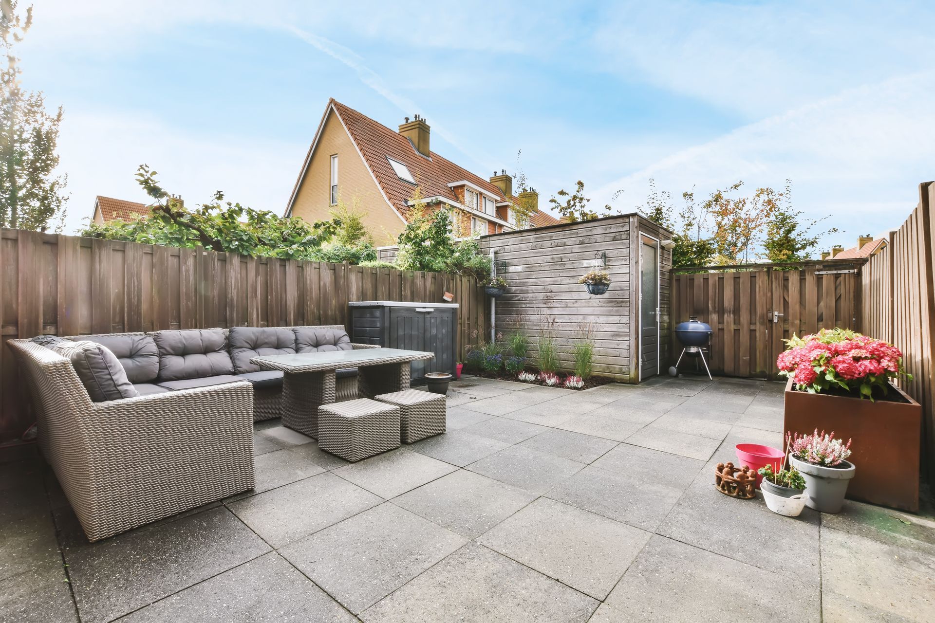 Patio with wicker seating, square stone tiles, wooden fence, and potted flowers in a sunny backyard