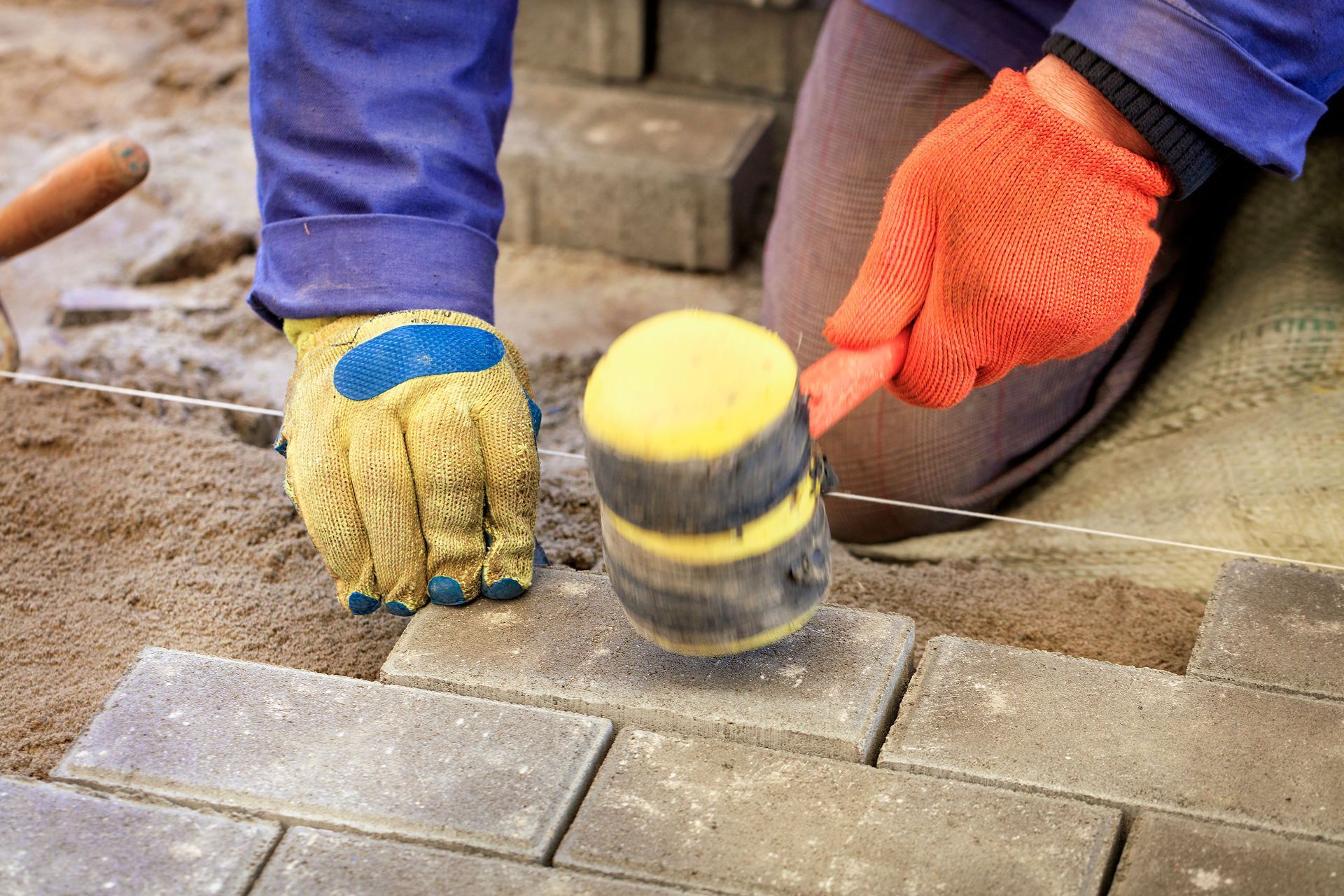 Hands in gloves laying brick pavers with a rubber mallet on a patio.