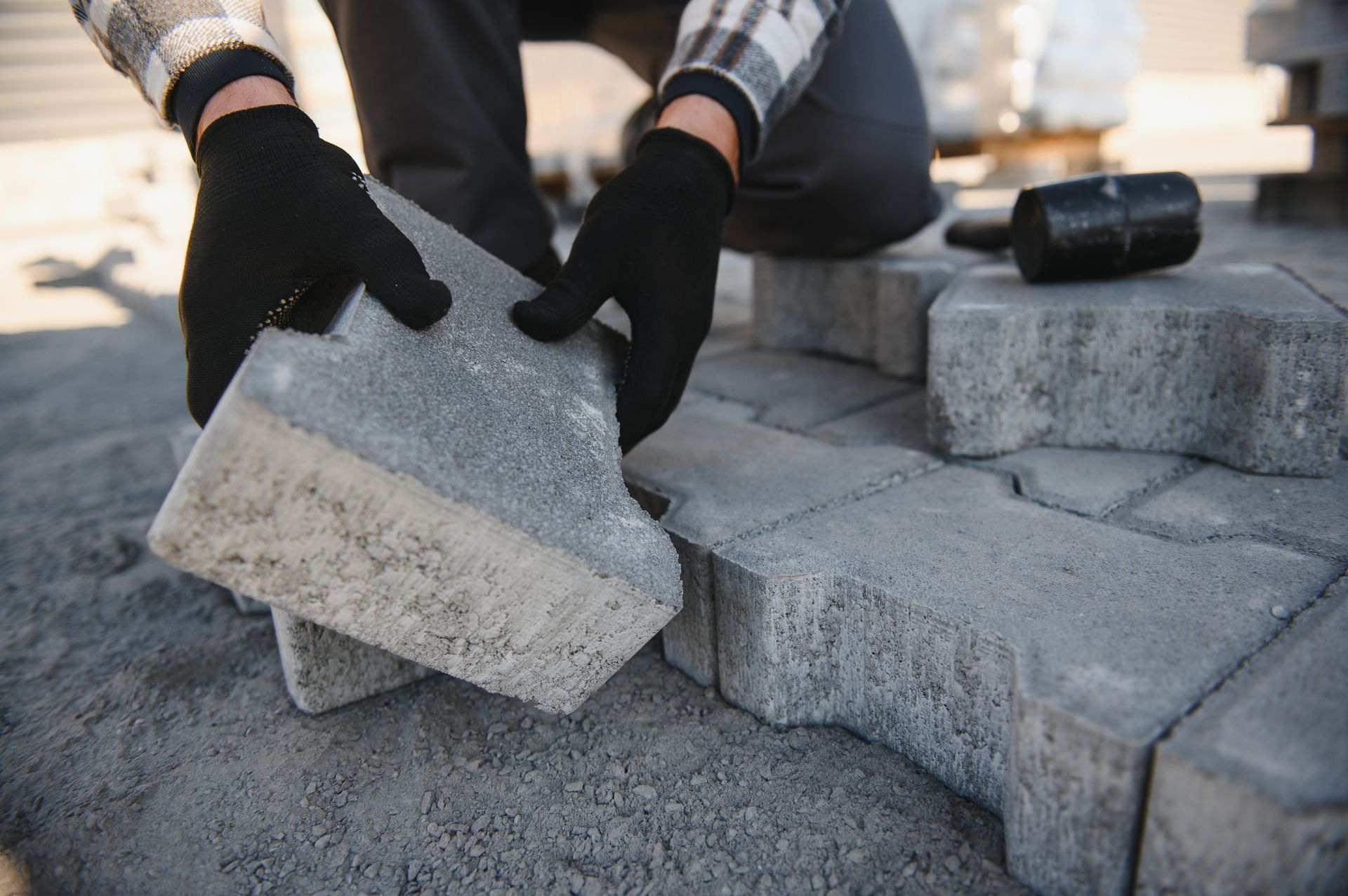 Gloved hands laying gray concrete pavers on a gravel surface outdoors