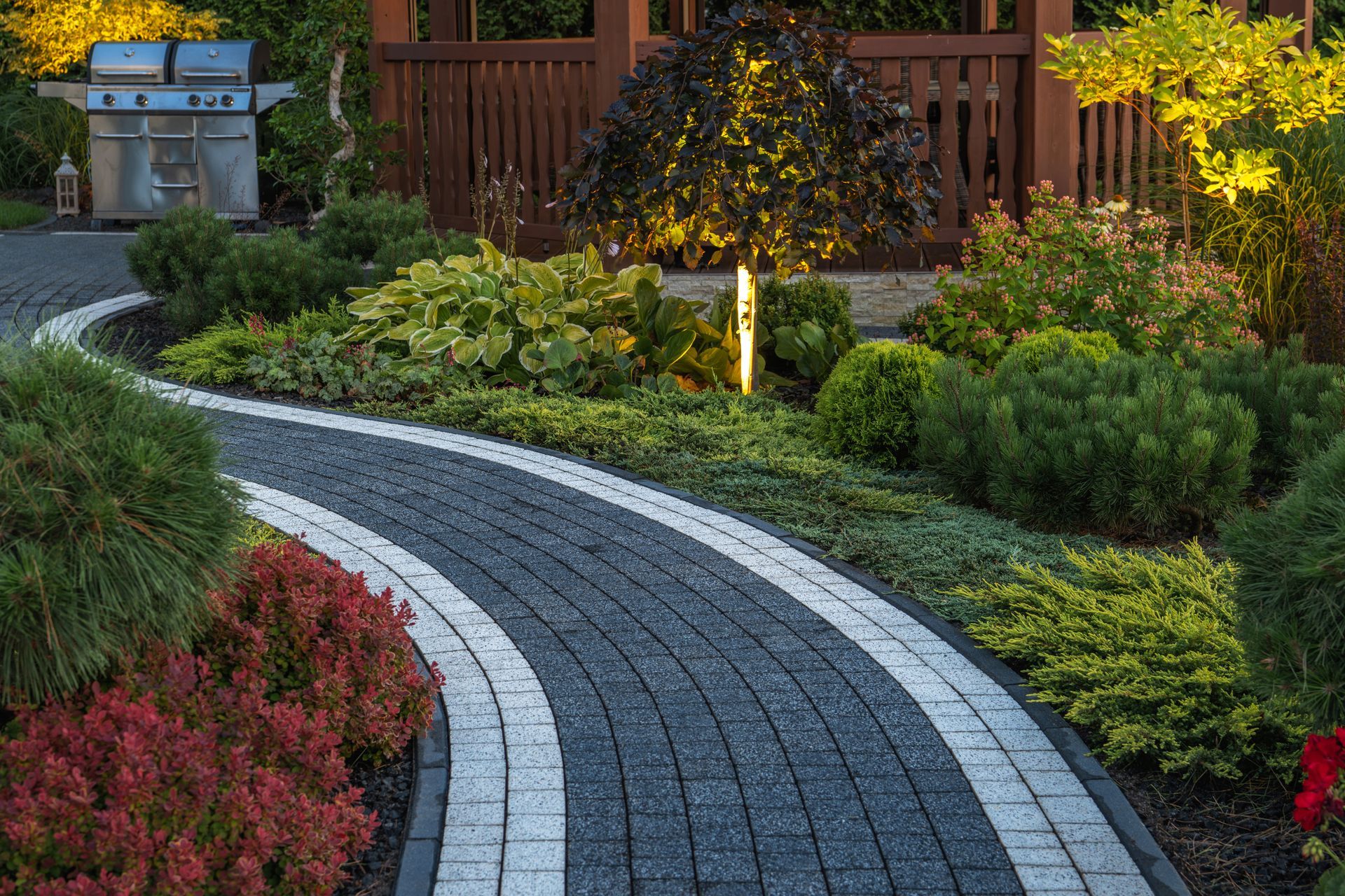 Curved stone garden path bordered by lush green and red shrubs, with a wooden fence in the background