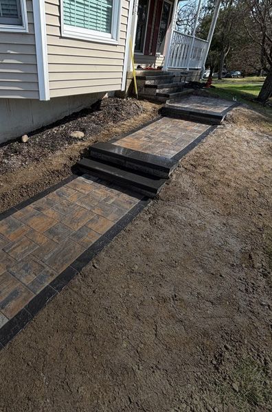 Paver walkway and steps leading to a house entrance. Brown and black pavers set in dirt.