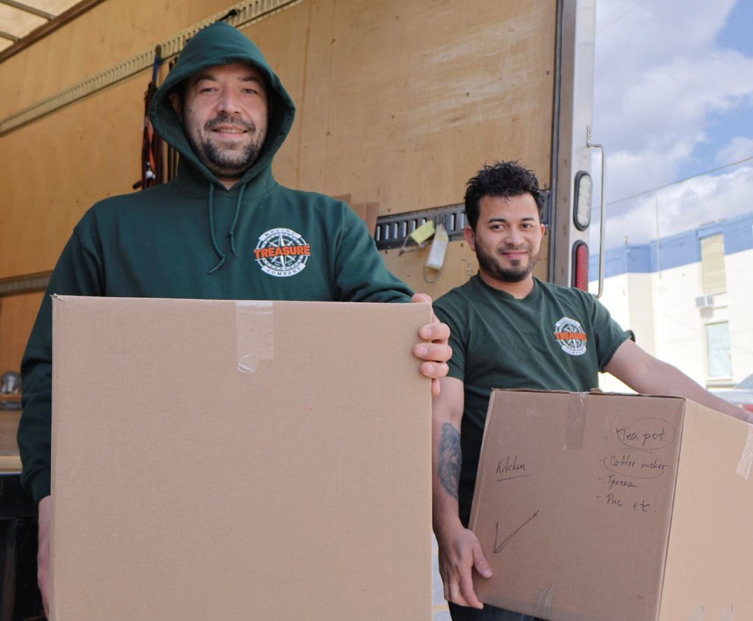 Two movers holding boxes smile, standing in a moving truck. One wears a green hoodie, the other a green shirt.