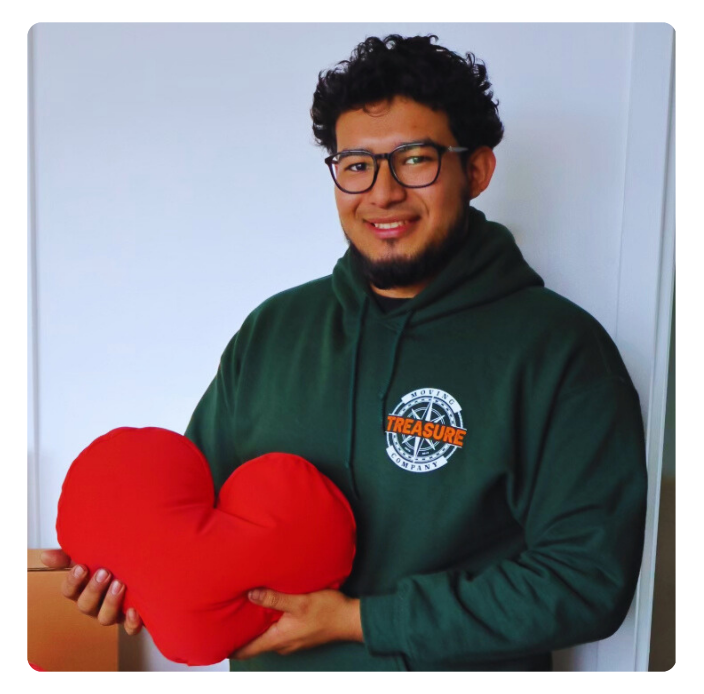 Man in green hoodie holding a large red heart-shaped pillow, smiling.