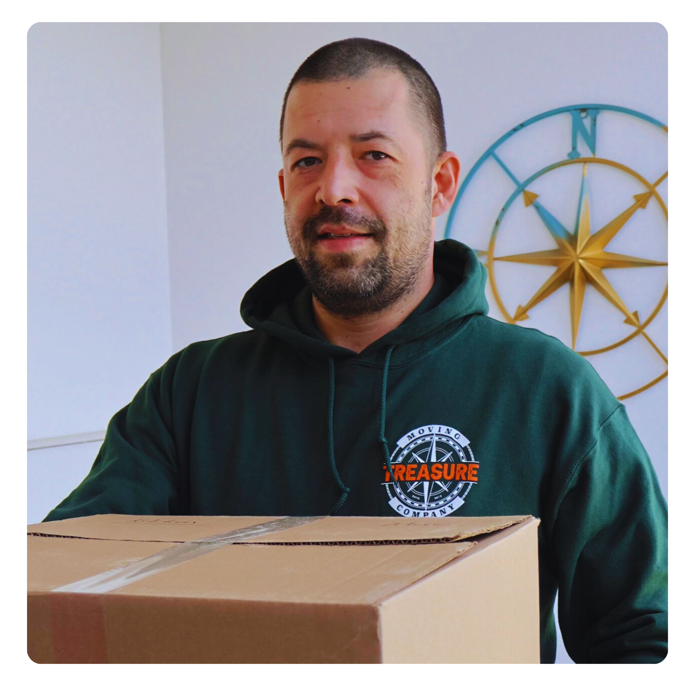 Man in green hoodie holding a cardboard box, with a nautical compass on the wall behind him.
