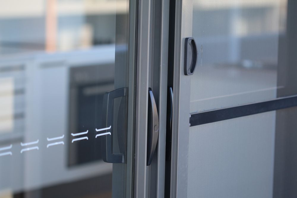 Sliding glass door with dark gray handles and frame, in a room with white walls and a view of a blurred kitchen. — Maryborough Bay Glass & Aluminium in Maryborough, QLD