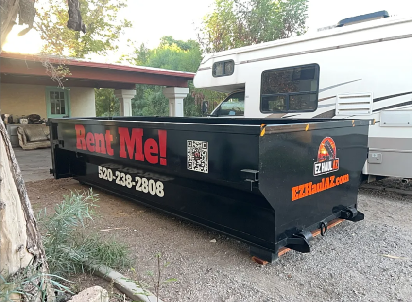 Black construction dumpster with “Rent Me!” text parked beside a white RV in a driveway.