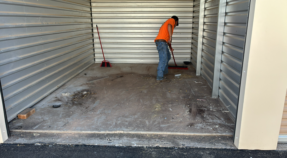 Worker in orange vest sweeping dirt inside an empty metal storage unit.