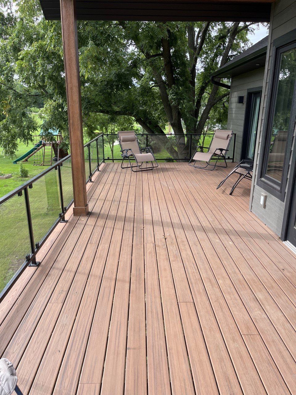 Covered wooden deck with two chairs and trees in the yard beyond.