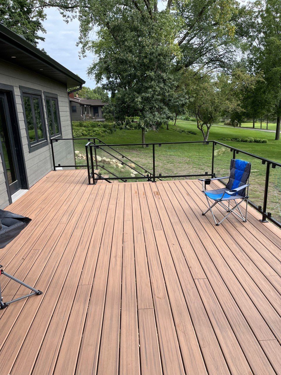 Wooden deck with black railing, blue folding chair, and grassy yard with trees beyond.