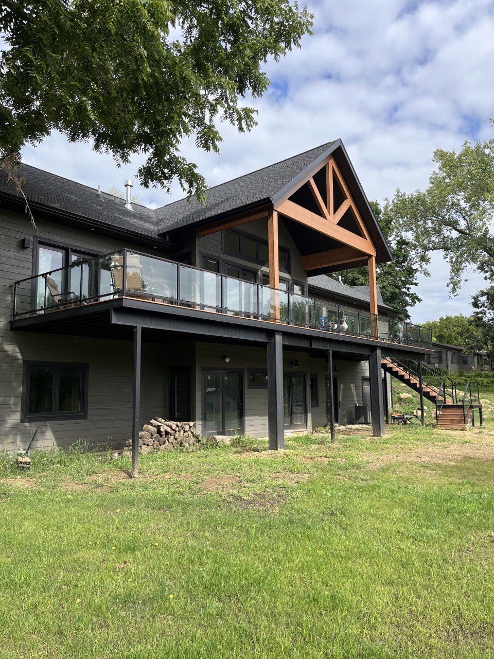 Modern two-story black house with a large deck and wood-trimmed gable, set on a grassy hillside under cloudy sky