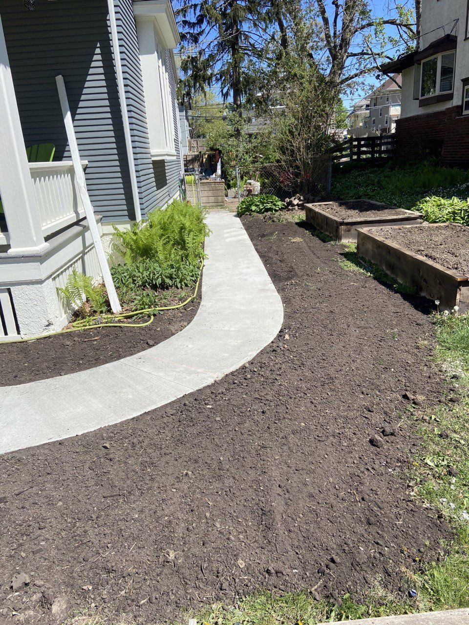 Curved concrete walkway beside a gray house, bordered by fresh soil and small shrubs in a residential yard.