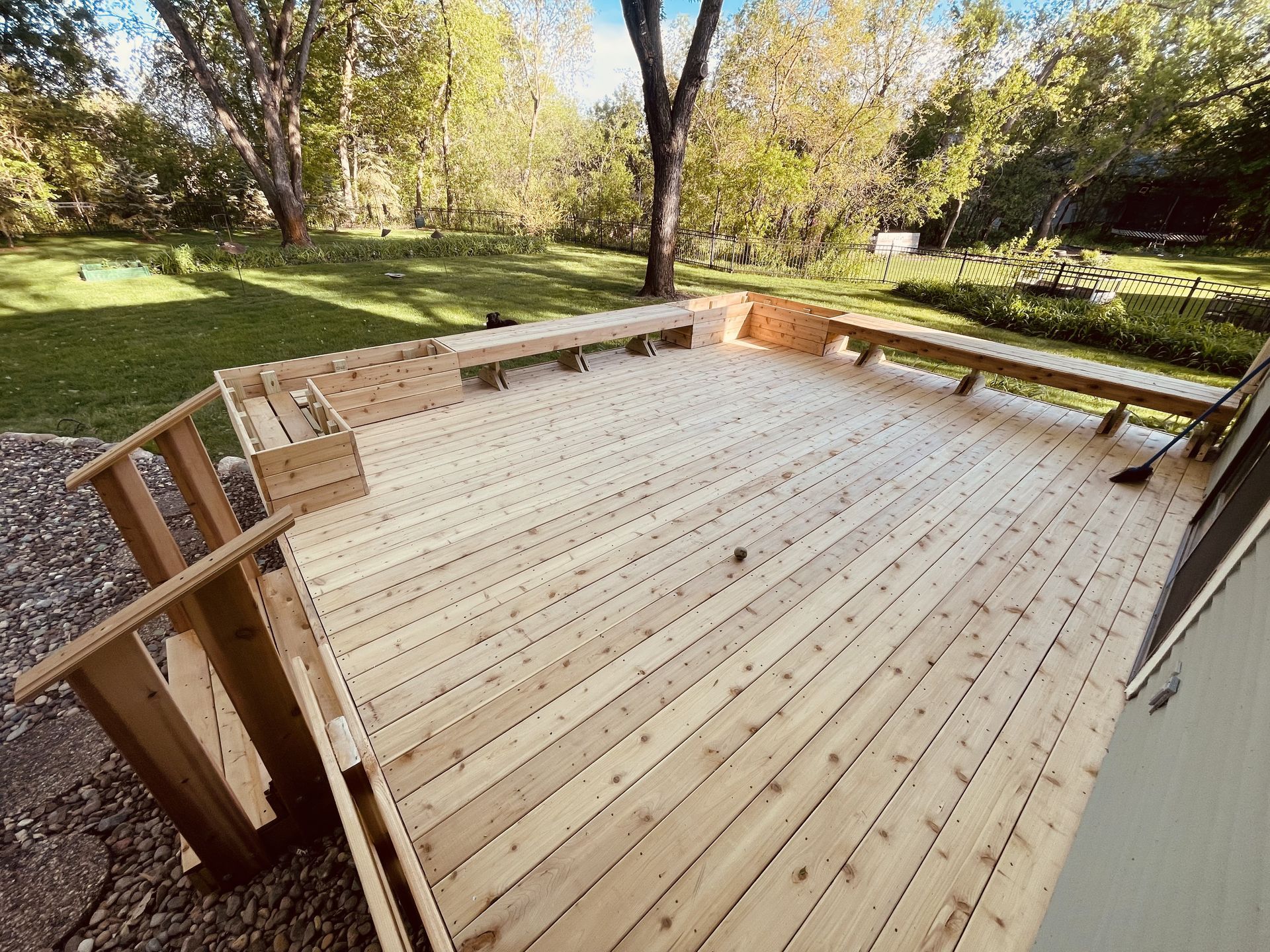 Wooden deck in a backyard setting with built-in benches, surrounded by greenery and trees.