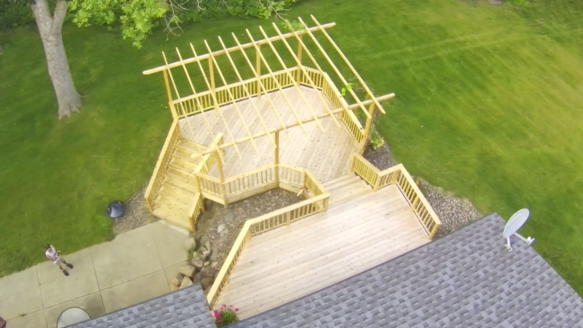 Aerial view of a wooden deck with stairs and overhead beams, next to a house on a grassy lawn.