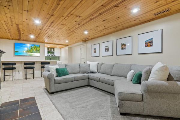 Cozy basement living space with a gray sectional, bar with stools, TV, and wood ceiling.