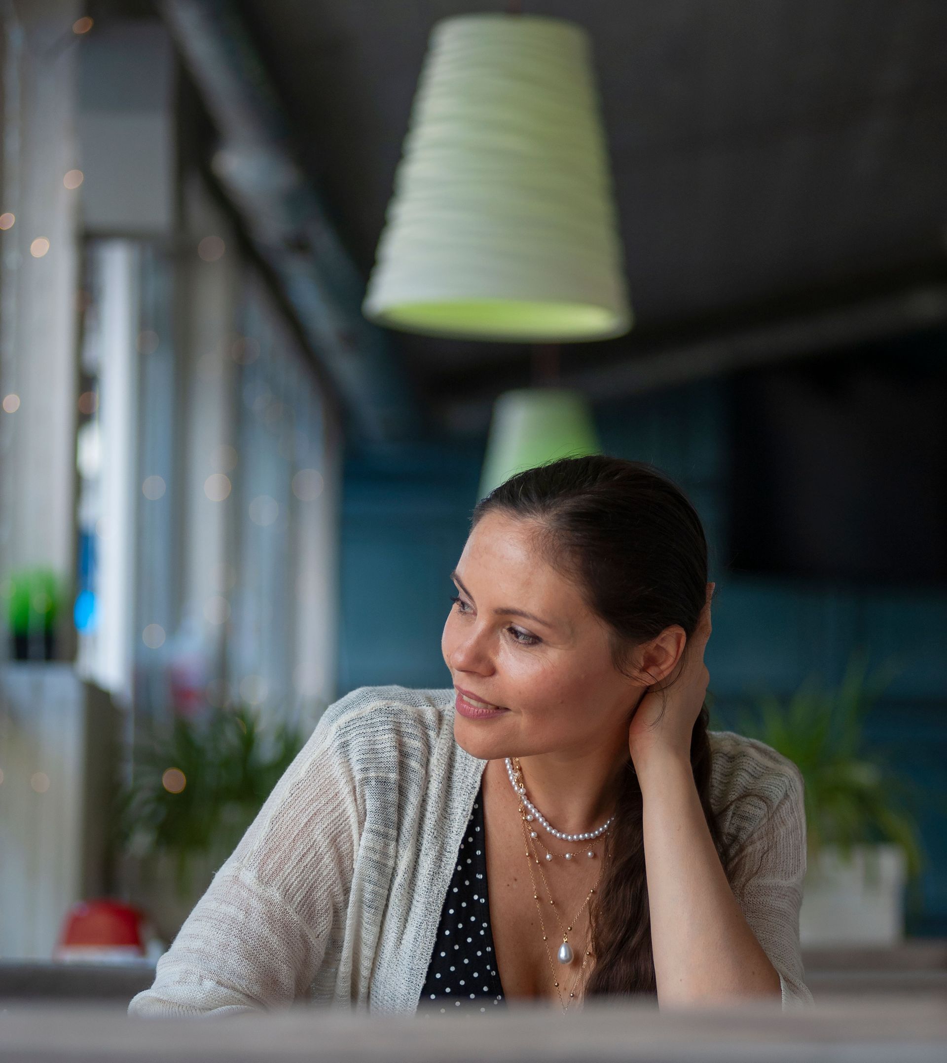 Woman seated at a café table, smiling and looking to the side under a hanging lamp.