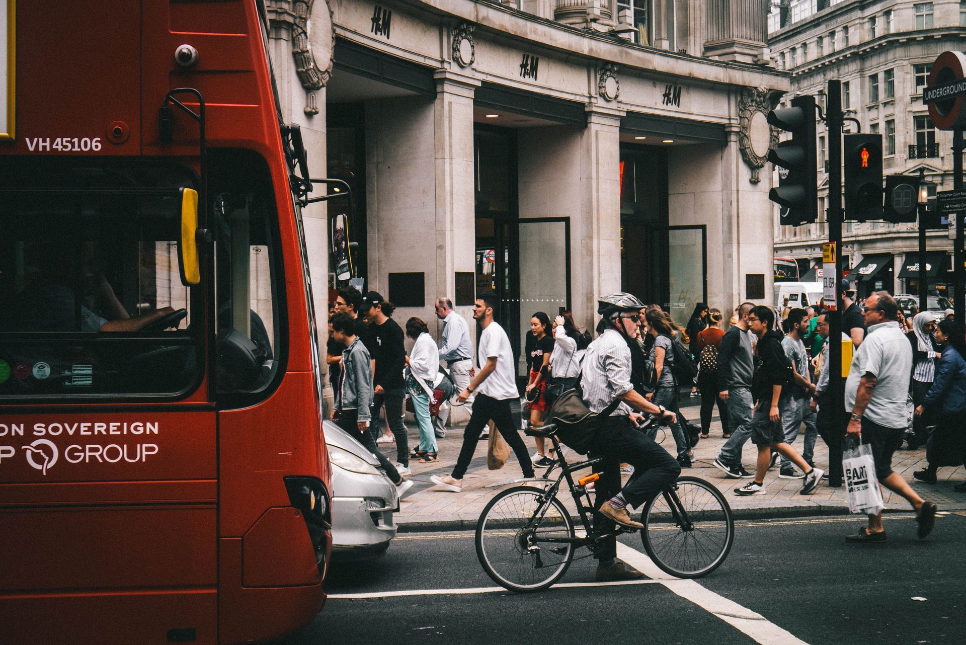 Crowded city street with pedestrians crossing beside a red bus and a cyclist in the foreground