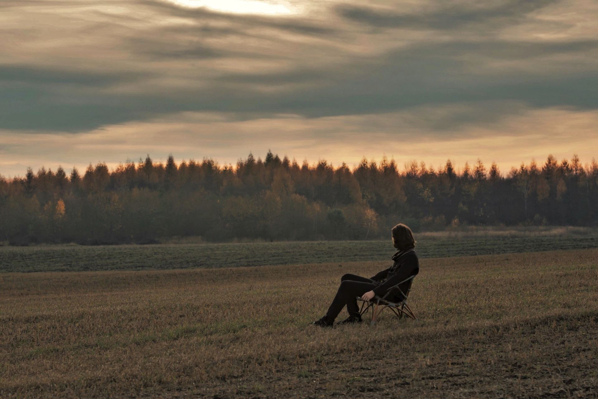 Person seated in a folding chair in a field at dusk, with a forest and cloudy sky in the background