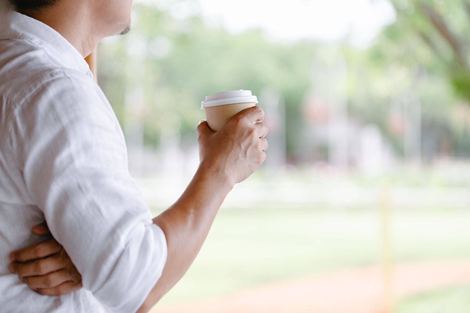 Person in a white shirt holding a coffee cup by a bright window.