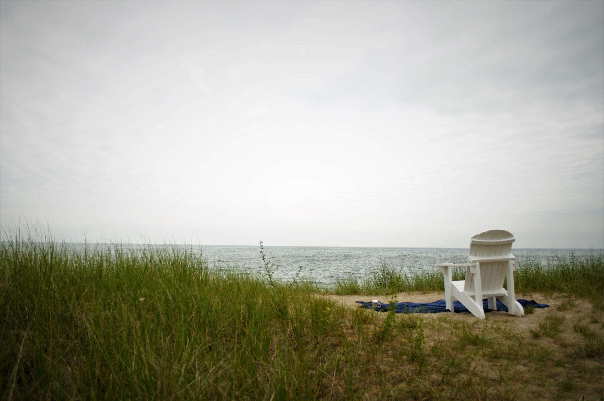 White beach chair on grassy dune by a calm gray ocean under an overcast sky